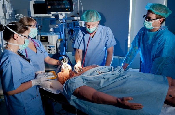 Bilde av fire medisinstudenter ved en sykehusseng. I sengen ligger en dukke, som de undersøker. Foto